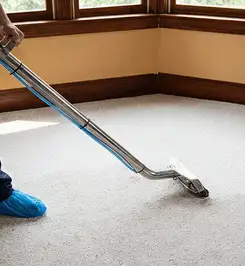 Steam carpet cleaning technician using a hot-water extraction wand on a residential carpet, wearing blue protective shoe covers Houston, TX.